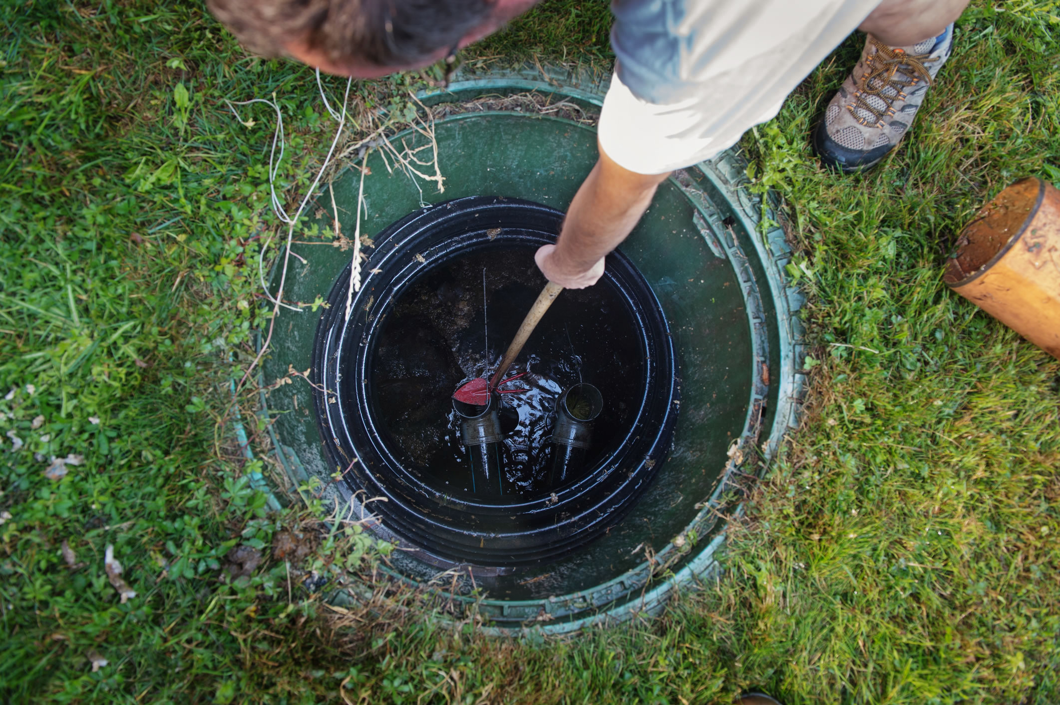 A person stands on grass and opens a green septic tank cover, inspecting or performing maintenance inside the tank with a tool in hand.