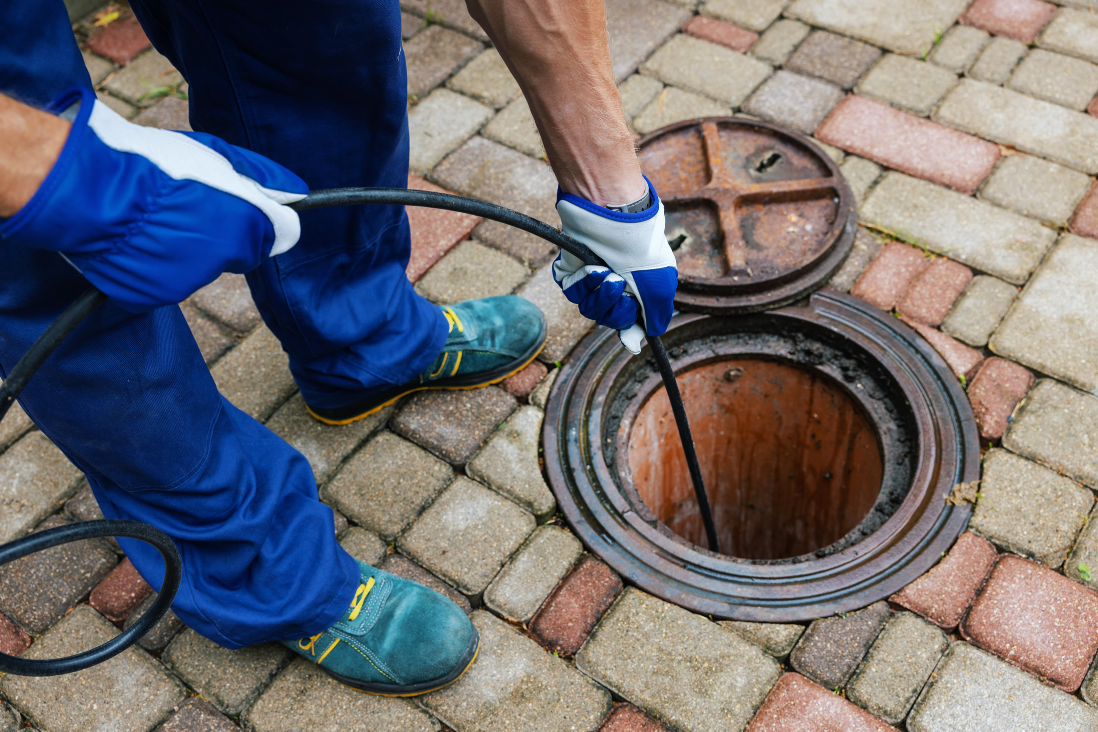 A person wearing blue gloves and work clothes uses a tool to clean or inspect a drain or sewer manhole on a tiled outdoor surface. The manhole cover is open, revealing the circular opening.