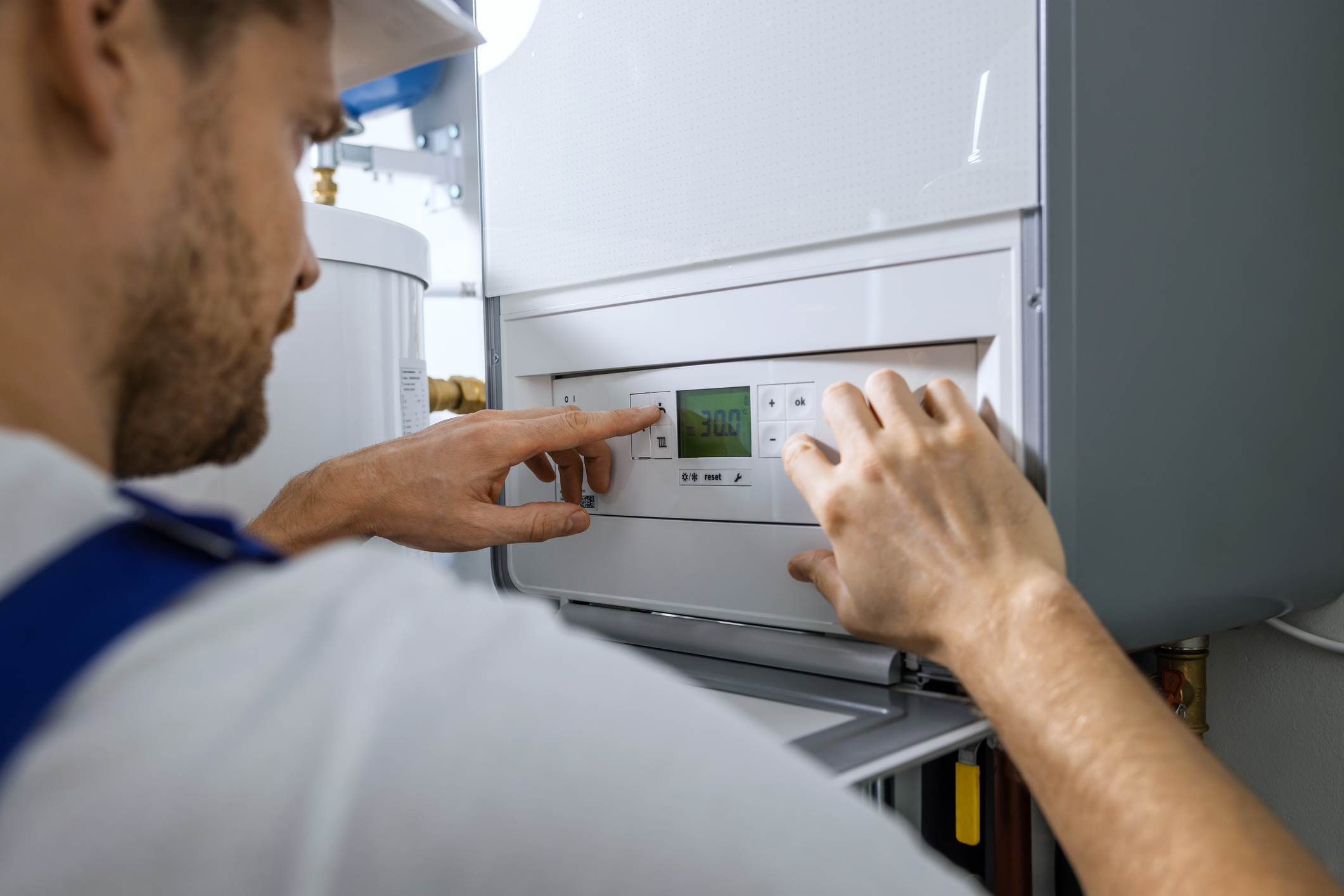 A person in a hard hat adjusts the settings on a wall-mounted digital control panel, possibly for a heating or HVAC system, inside a utility room.