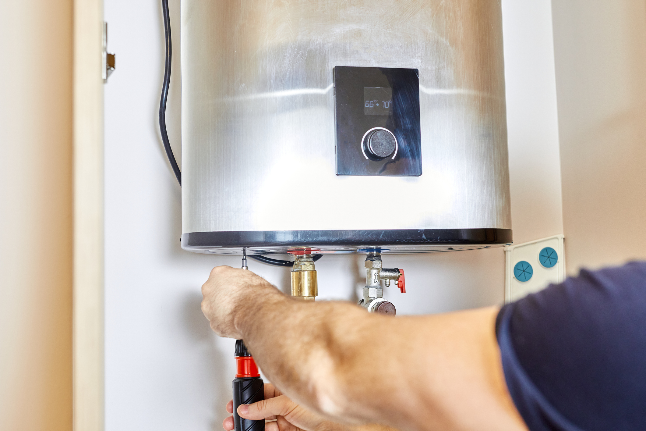 A person uses a screwdriver to adjust or repair a wall-mounted water heater. The water heater has a digital display and is installed indoors, with pipes and wires connected underneath.