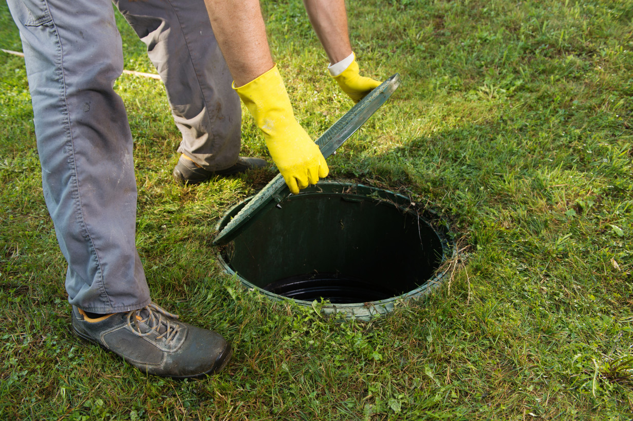 A person wearing yellow gloves and gray pants lifts the lid of an outdoor septic tank on a grassy lawn.
